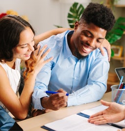 Young smiling coupl sign a housing document at a realtor's desk