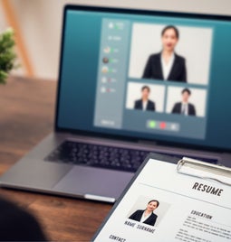 A clipboard with a paper resume in front of a laptop screen with professional headshots on the screen.