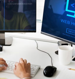 Person taps on keyboard in front of two desktop screens looking at web hosting