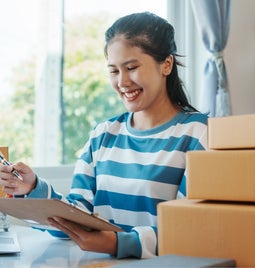 Woman stands behind a pile of package boxes smiling at her clipboard