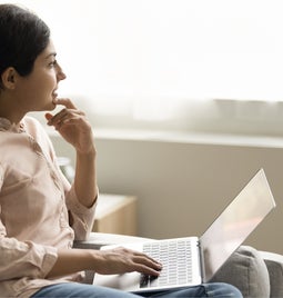 Young woman sits on sofa holding laptop looking away throughtfully