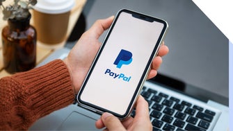 Person holds a phone showing the PayPal logo with a laptop in the background