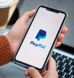 Person holds a phone showing the PayPal logo with a laptop in the background