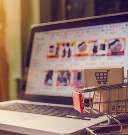 A small shopping cart with cardboard boxes positioned in front of a laptop with an online store on the screen.