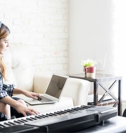 A musician with headphones on, with one hand on her laptop and another on a keyboard.