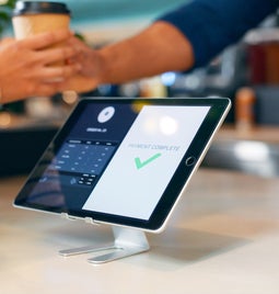 Seller hands customer a coffee behind a POS tablet showing message 