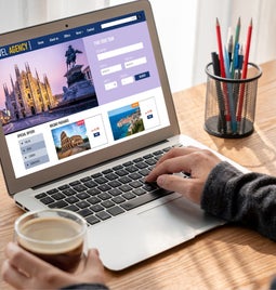 Screenshot of a man using a laptop with a travel bookings website on screen.